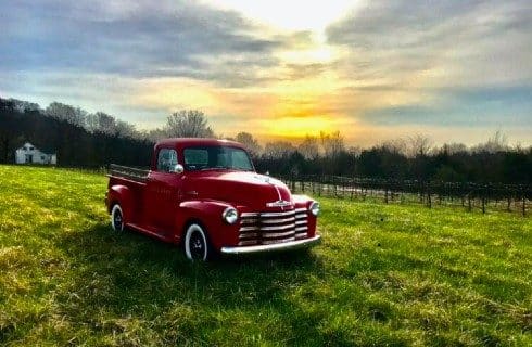 Bright red vintage pick up truck in a grassy field with sun and clouds overhead