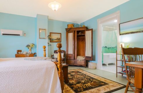 Bedroom with queen bed with white quilt, tall wooden armoire and doorway into bathroom with white clawfoot tub