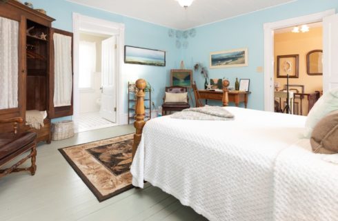 Large bedroom with blue walls, queen bed, armoire, writing desk and doorway into a white bathroom