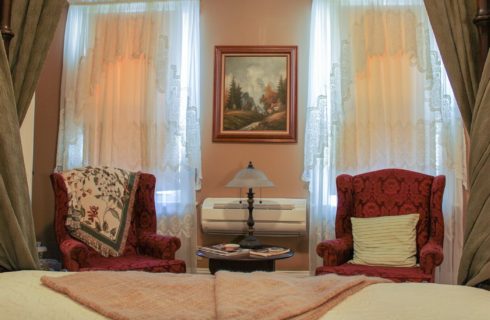 Two red wingback sitting chairs, each in front of a large window with sheer lacy white curtains