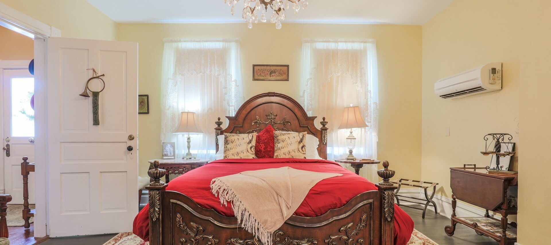 Bedroom with queen bed, antique headboard, red linens and two windows with white sheer curtains