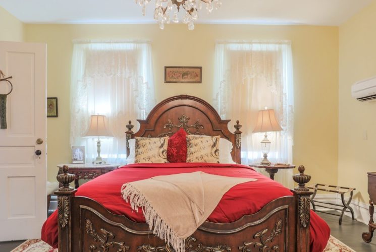 Bedroom with queen bed, antique headboard, red linens and two windows with white sheer curtains