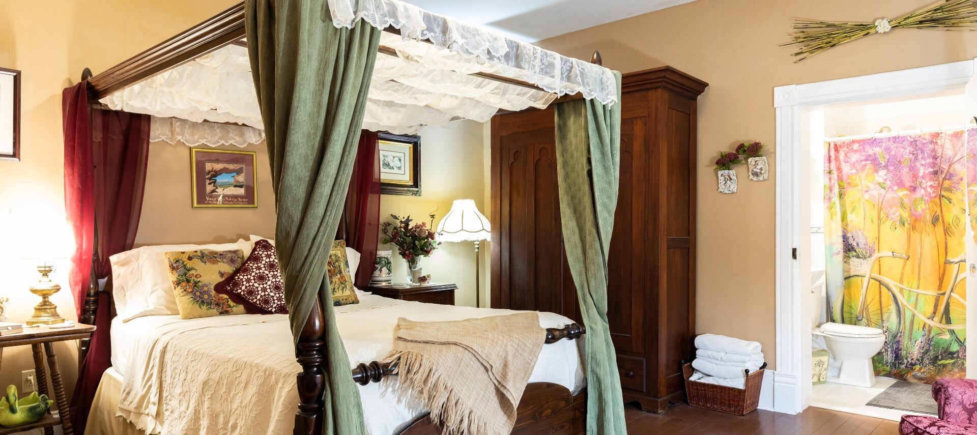 Bedroom with large four poster canopy bed. tall brown armoire and doorway into a bathroom with colorful shower curtain