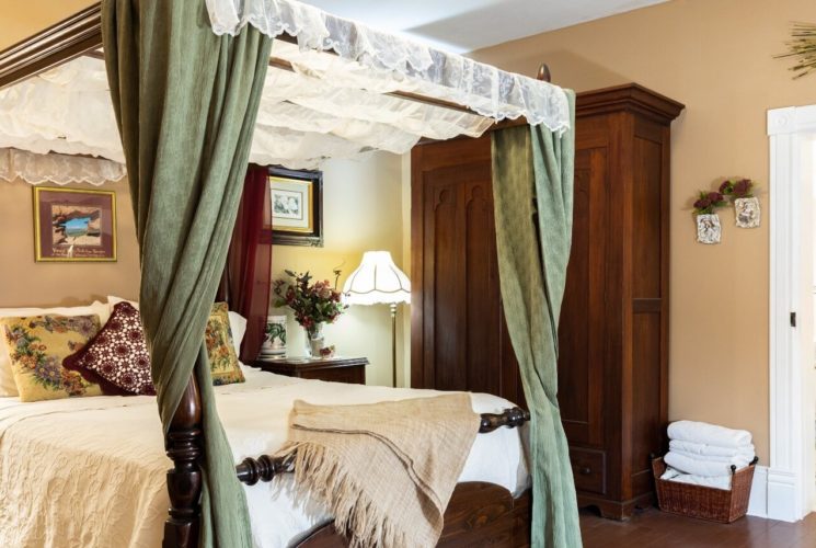 Bedroom with large four poster canopy bed. tall brown armoire and doorway into a bathroom with colorful shower curtain