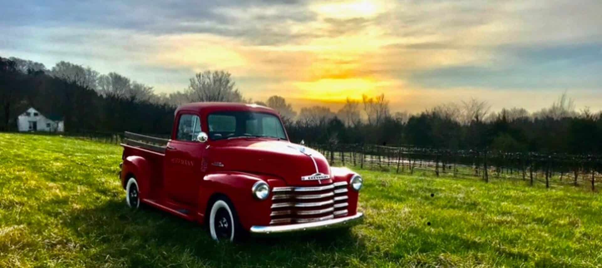 Bright red vintage pick up truck in a grassy field with sun and clouds overhead